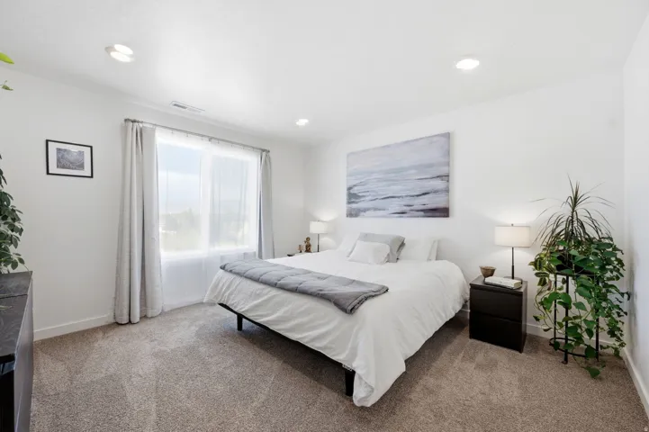 Bright bedroom featuring neutral-tone carpeting, a large window with privacy sheers, recessed ceiling lighting, and crisp white walls