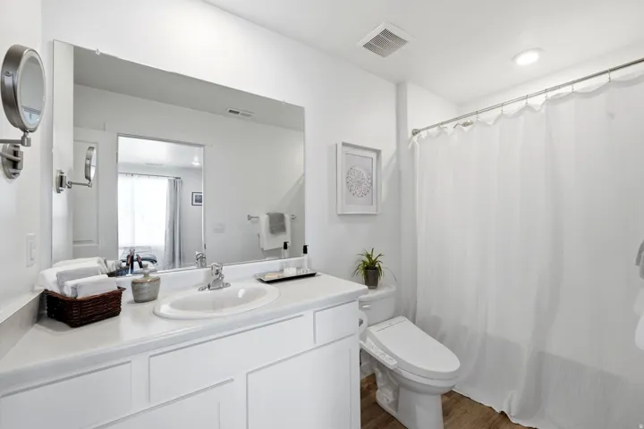 Bathroom featuring a single vanity with an integrated sink, a large wall-mounted mirror, wood-finish flooring, a commode, and a shower with a privacy curtain