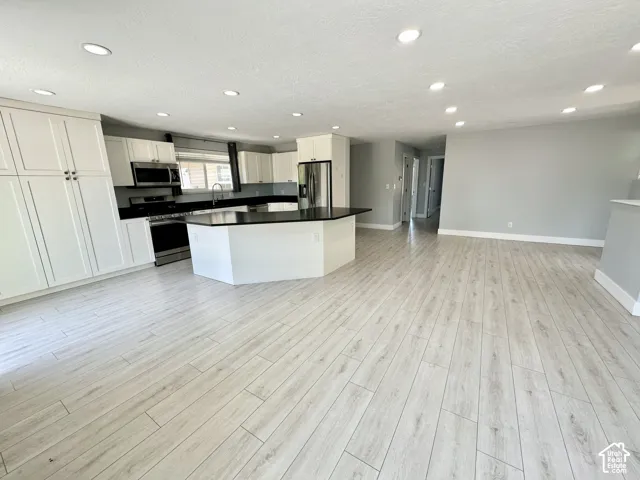 Spacious kitchen featuring an angular island with a dark countertop, white shaker cabinetry, stainless steel appliances, and light wood-finish flooring