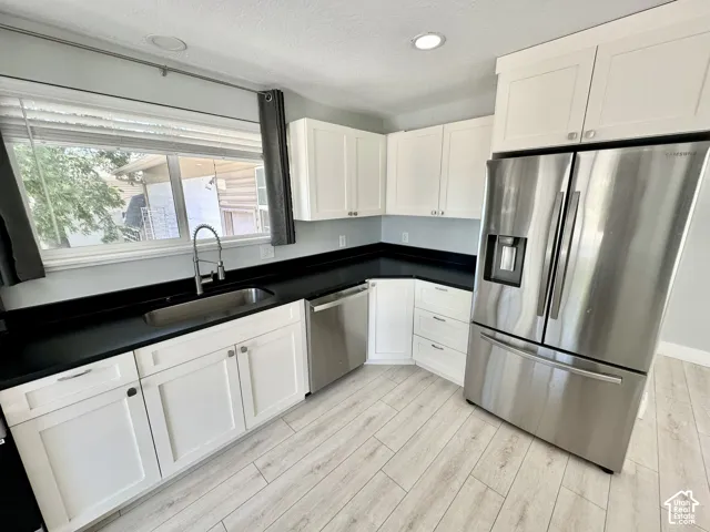 Kitchen featuring white shaker cabinetry, dark countertops, stainless steel appliances, a stainless steel sink with a pull-down faucet, and light wood-finish flooring