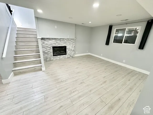 Spacious room featuring light wood-finish flooring, a contemporary fireplace with a stacked stone facade and white mantel, and a staircase with matching wood-finish treads and white risers