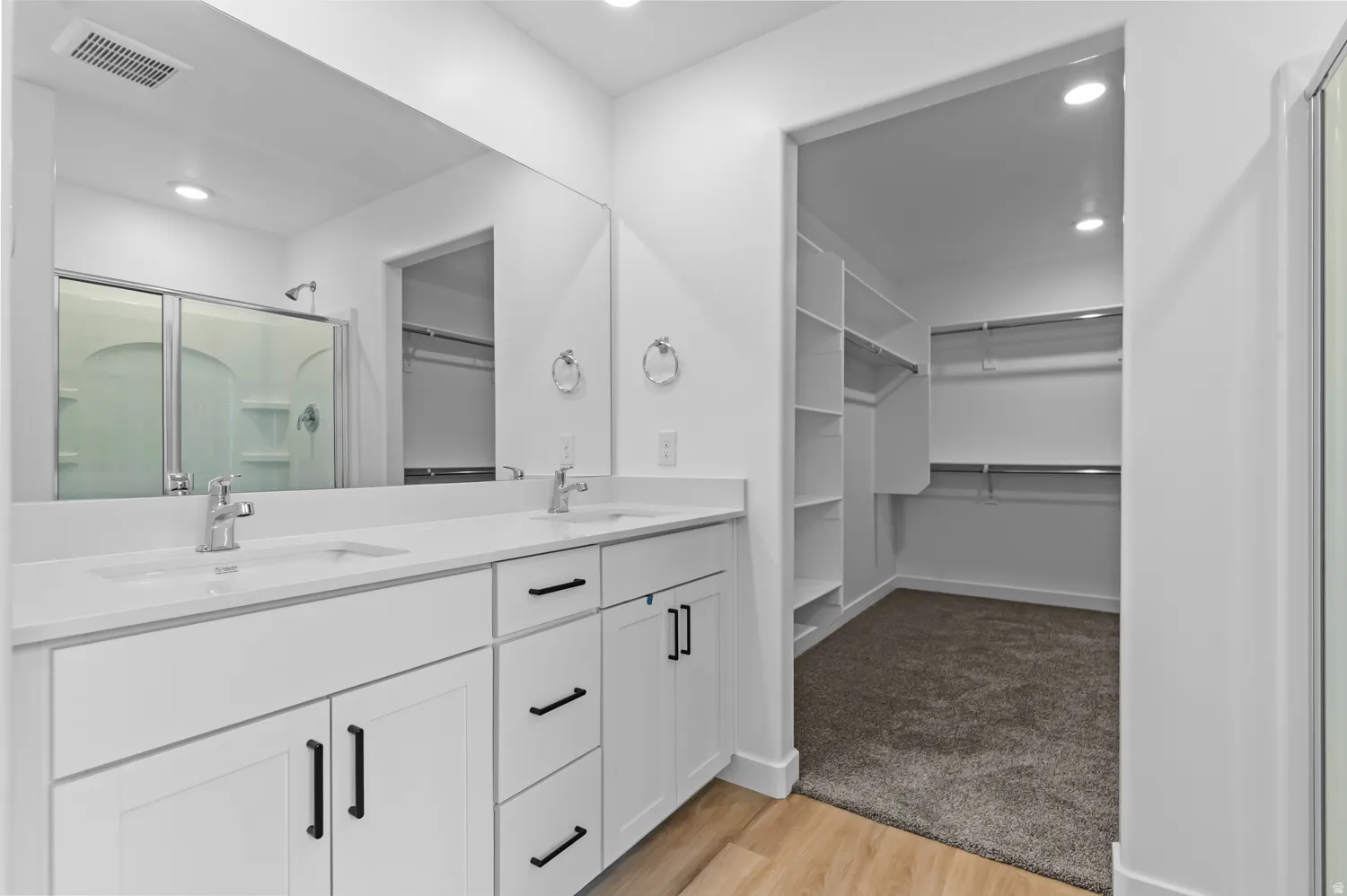 Dual vanity with white cabinetry and dark hardware, large wall-mounted mirror, and a walk-in closet featuring built-in shelving and hanging rods