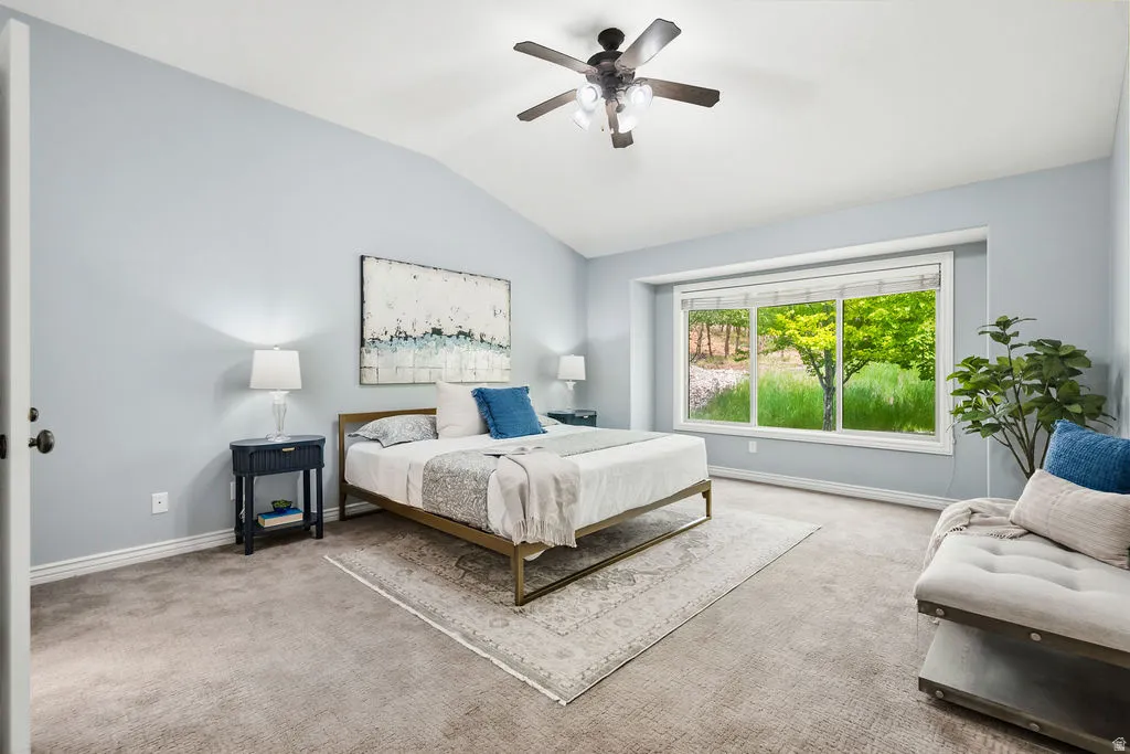 Vaulted ceiling bedroom featuring a large picture window, ceiling fan, and carpeted flooring