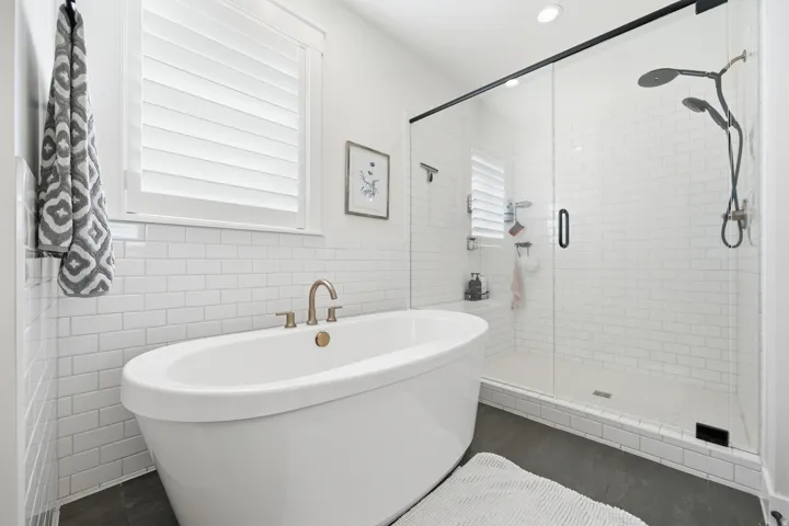 Freestanding soaking tub with brushed nickel fixtures, glass-enclosed shower with black trim, dual shower heads, white subway tile surround, and dark wood-finish flooring