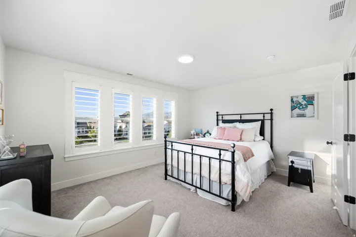 Bright bedroom featuring multiple windows with louvered shutters, light gray carpet flooring, white walls, and a recessed ceiling light