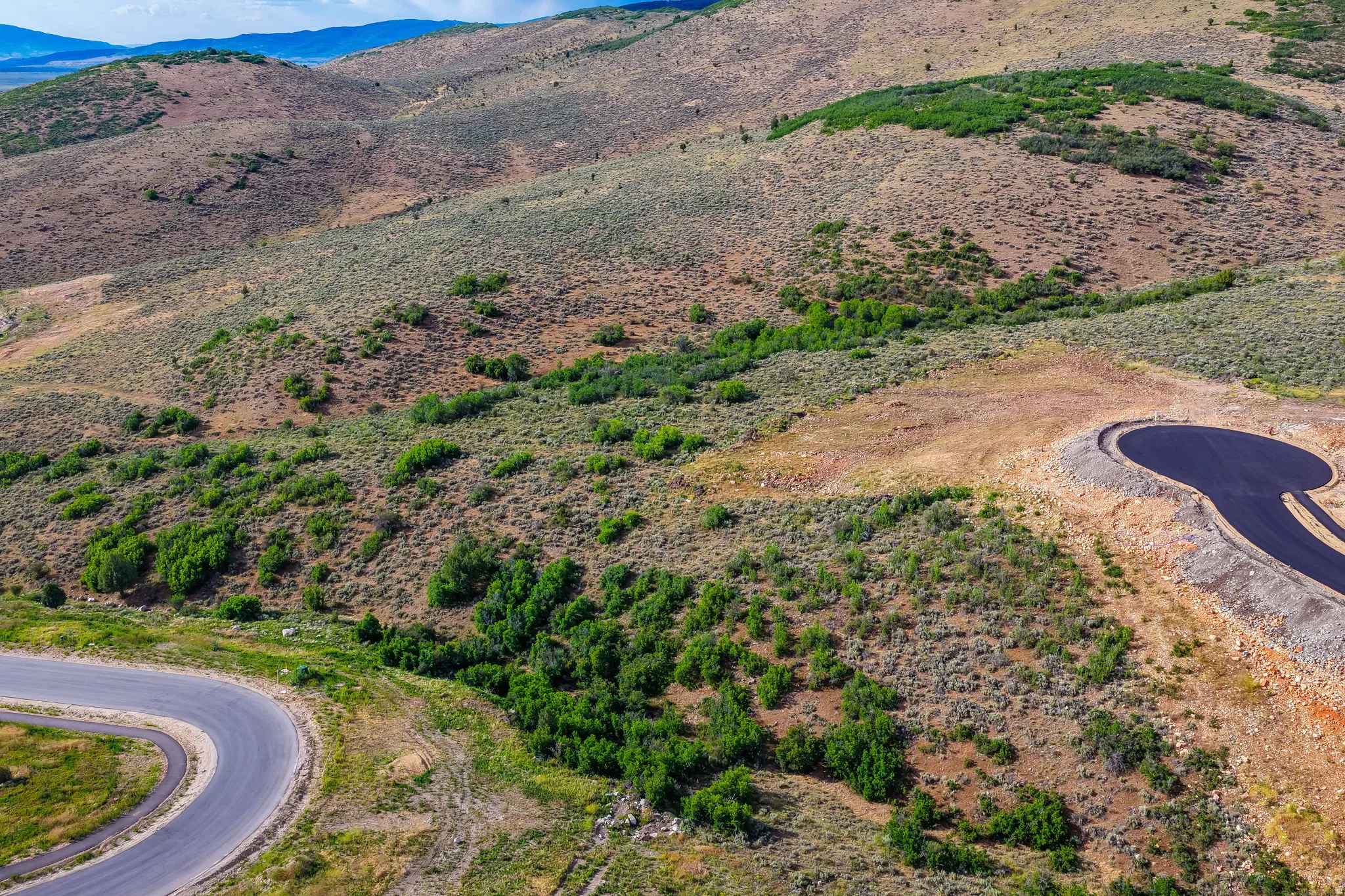 Expansive mountain terrain featuring natural vegetation, a newly paved cul-de-sac, and a winding paved road