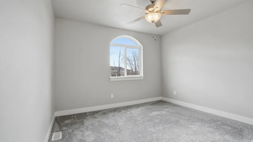 Spacious room featuring an arched window, a ceiling fan with integrated lighting, light gray wall paint, and gray textured carpeting with white baseboards