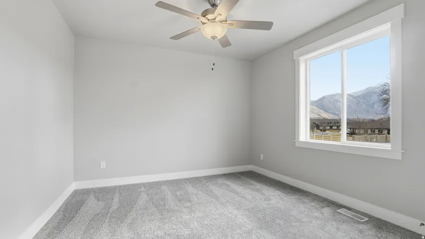 Spacious room featuring light gray walls, gray patterned carpeting, a white trim window, and a ceiling fan with integrated lighting