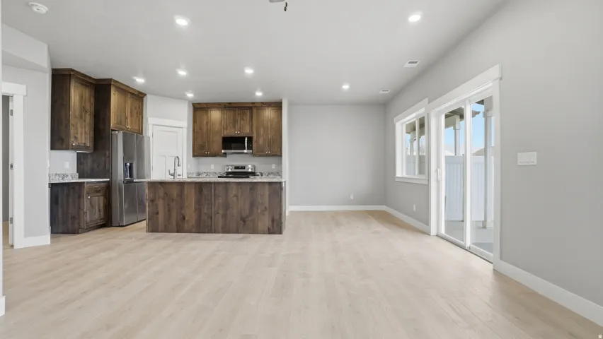 Open-concept living area featuring light wood-finish flooring, recessed lighting, and a modern kitchen with dark wood cabinetry, stainless steel appliances, and a central island with a sink