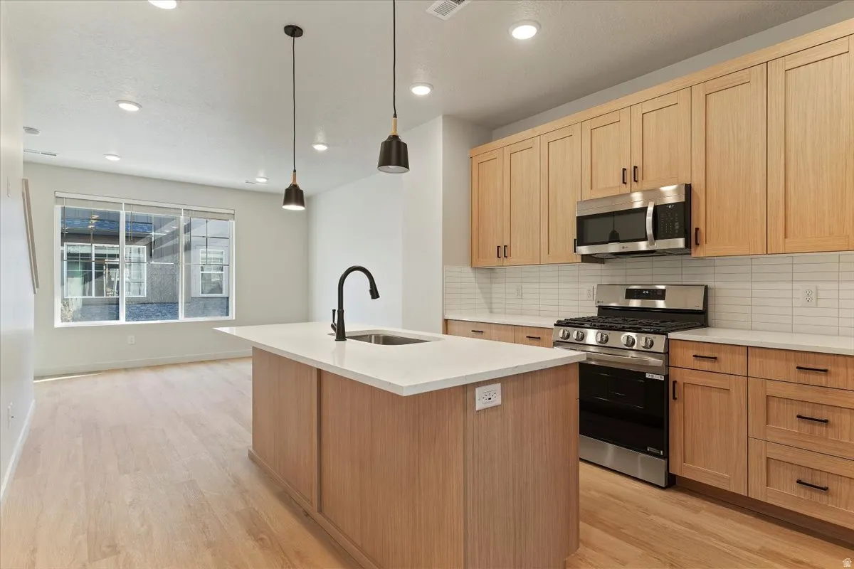 Open concept kitchen featuring an island with a white countertop and undermount sink, pendant lighting, stainless steel appliances, light wood-finish cabinetry, and a horizontal tile backsplash