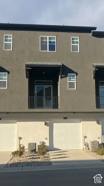 Multi-story exterior featuring a stucco facade, private balcony with metal railing, single-car garage, and low-maintenance rock landscaping