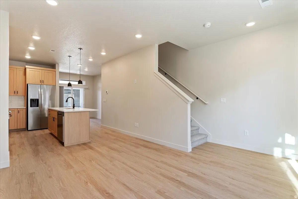 Open-concept living area featuring light wood-finish flooring, a kitchen island with pendant lighting, stainless steel appliances, and a carpeted staircase with white railings