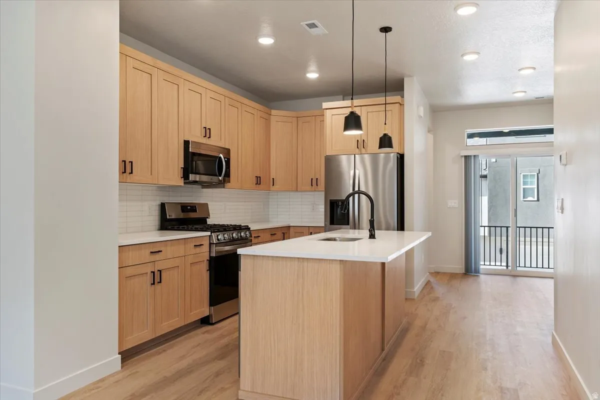Open-concept kitchen featuring wood-finish cabinetry, stainless steel appliances, white subway tile backsplash, and an island with a matte black faucet