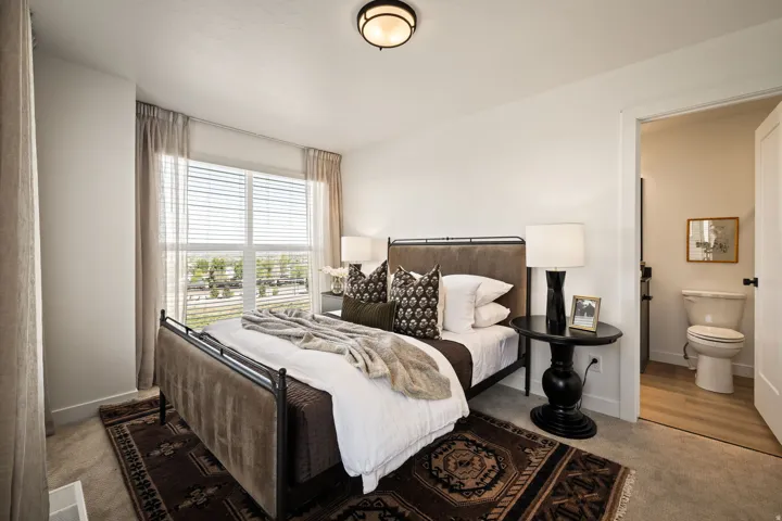 Carpeted bedroom featuring a large window with blinds and drapes, an overhead light fixture, and an entry to a bathroom with wood-finish flooring