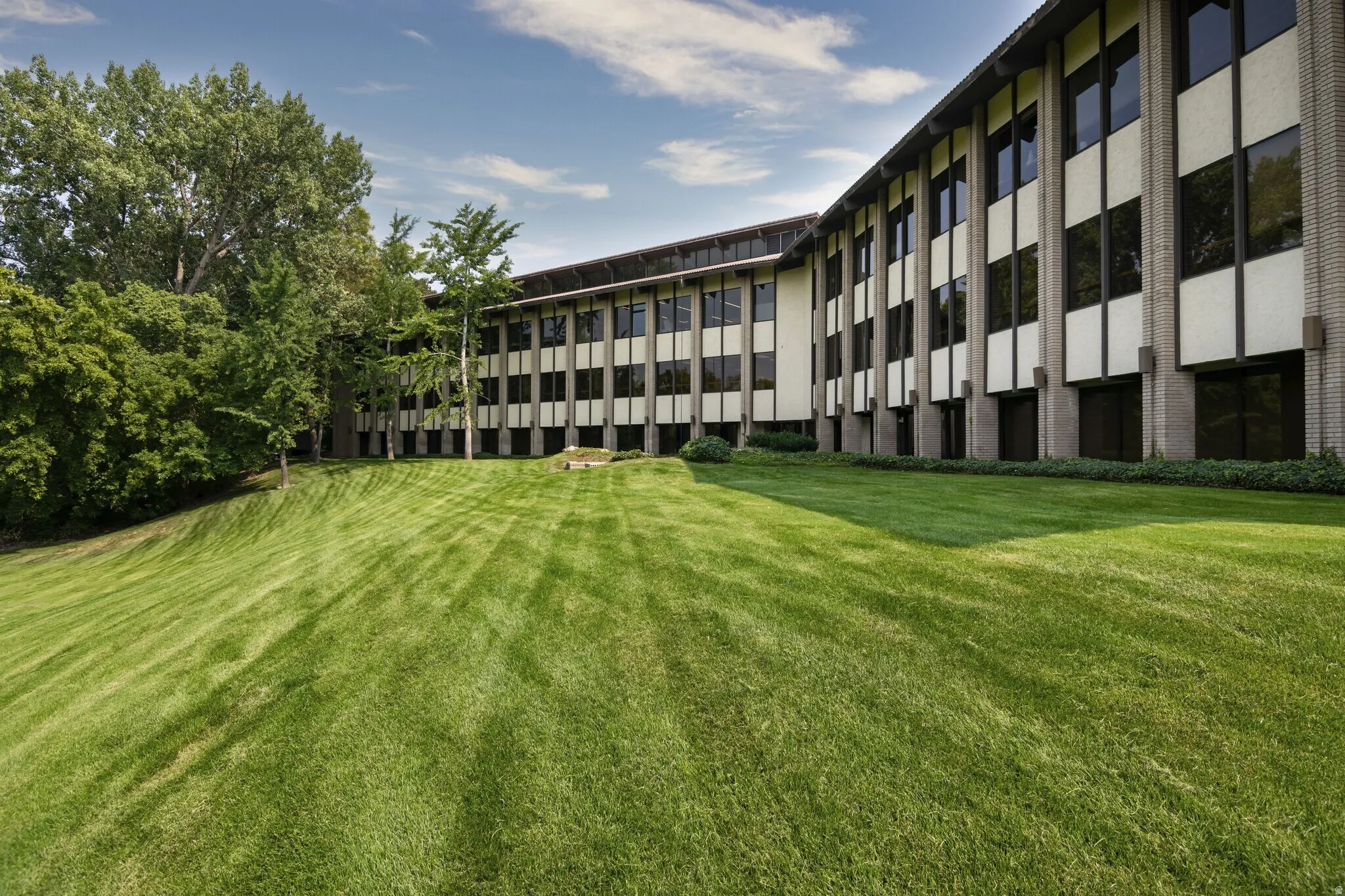 Expansive green lawn sloping towards a multi-story building featuring dark-framed windows, light-colored panels, and brick accents