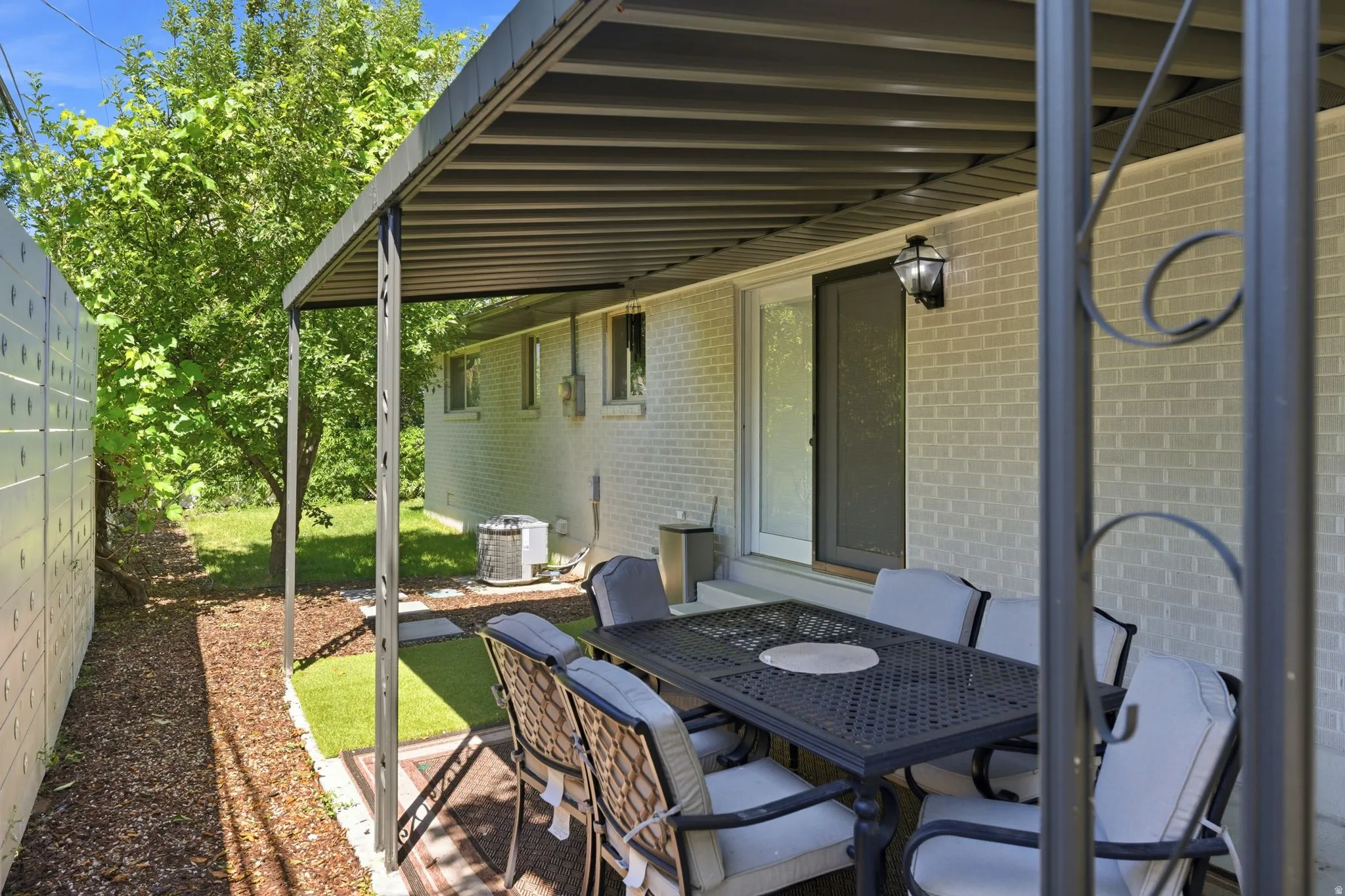 Covered patio area featuring a metal pergola, light-colored brick exterior, sliding glass door, exterior wall lantern, and partial artificial turf surfacing