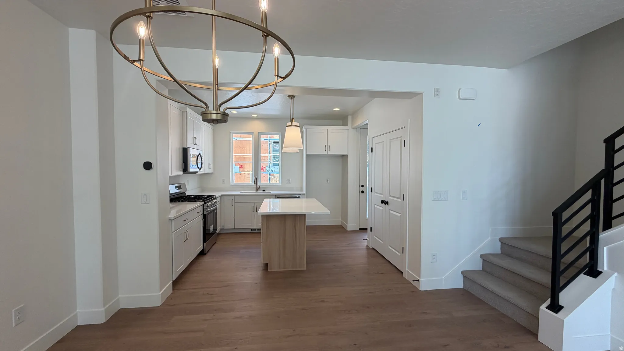 Open-concept kitchen and living area featuring white cabinetry, stainless steel appliances, a light wood-finish flooring, a contemporary chandelier, and a staircase with black metal railings