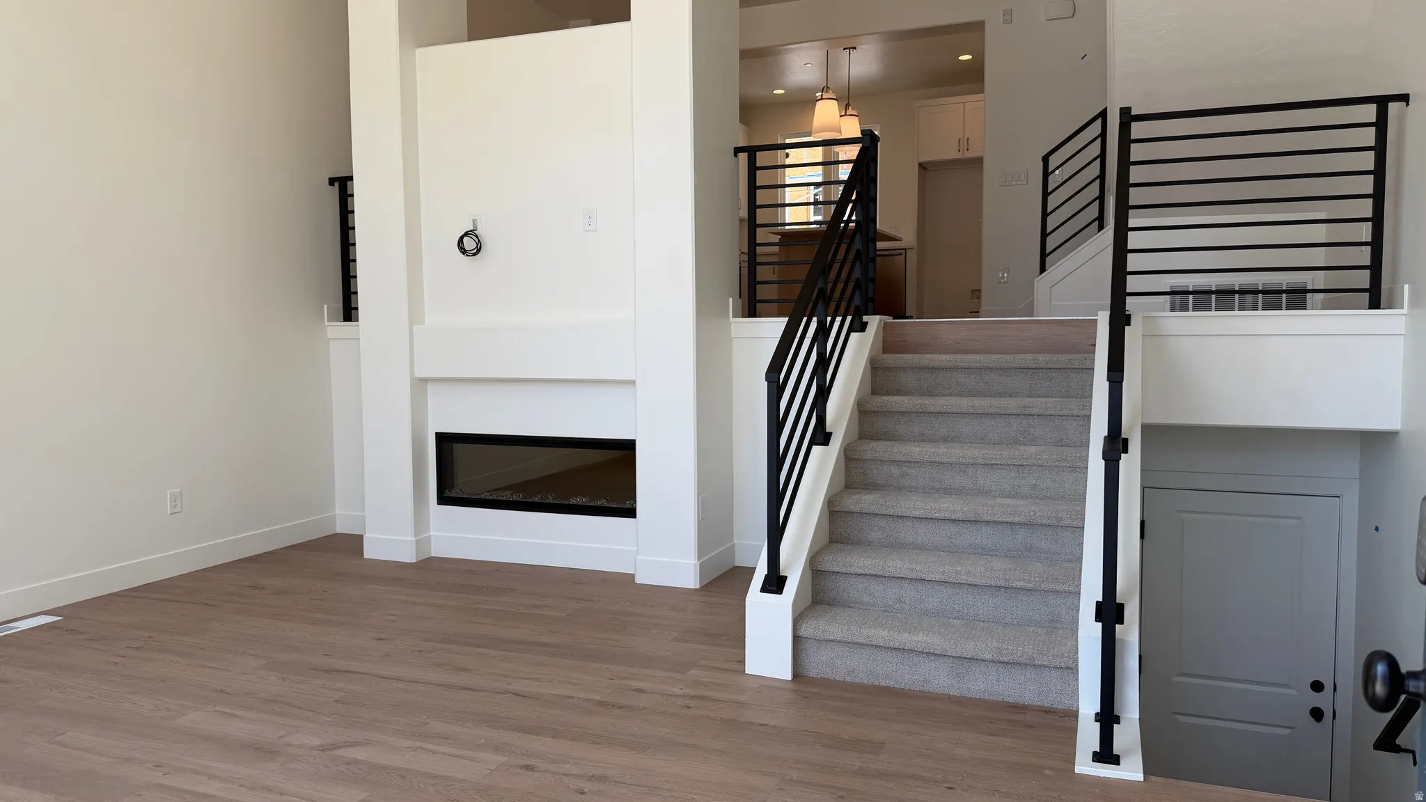 Open-concept interior featuring wood-finish flooring, a modern fireplace, carpeted staircase with contemporary black metal railings, and a lower-level access door