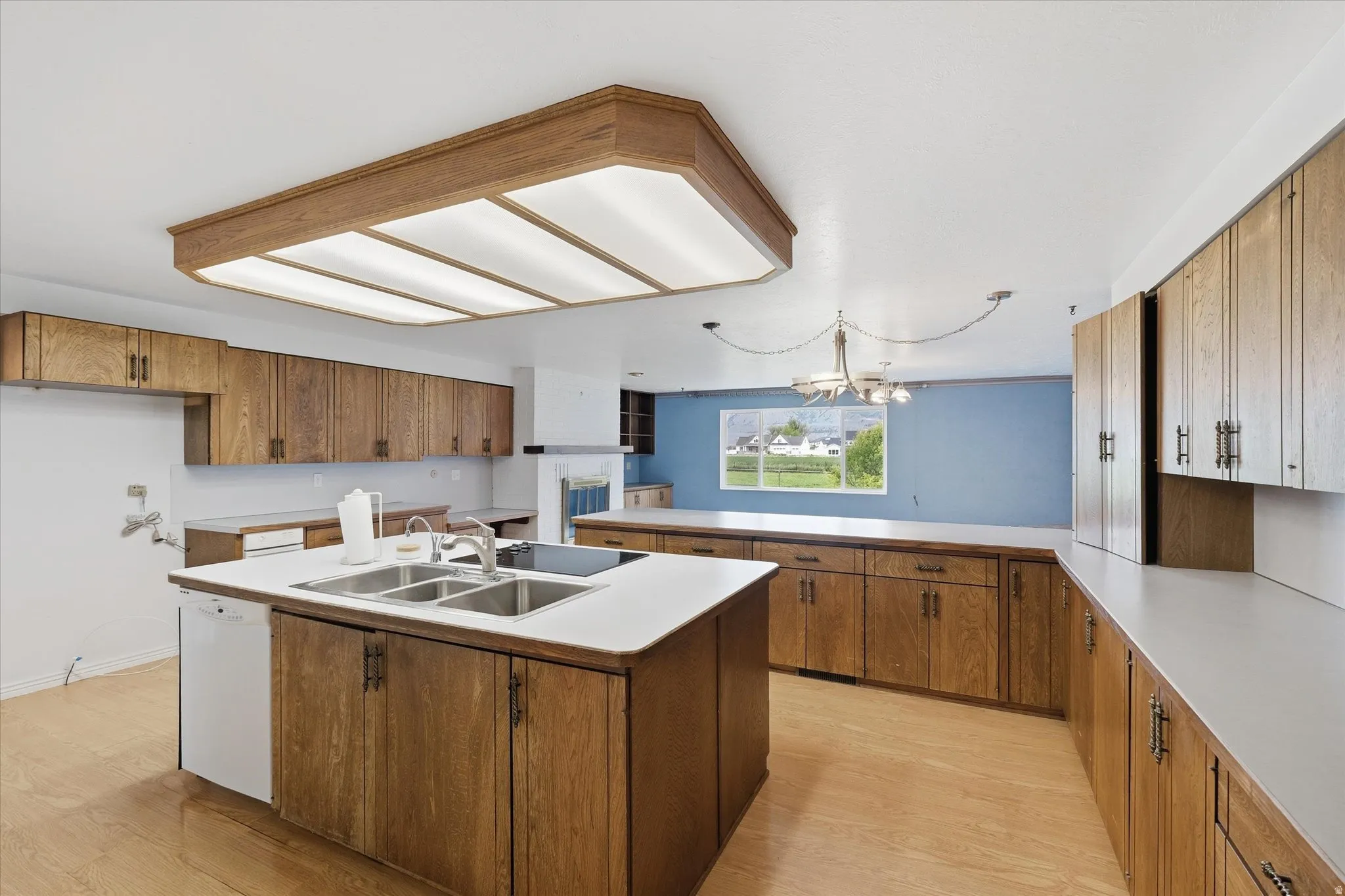 Kitchen featuring wood-finish cabinetry, extensive countertop space, a central island with an integrated sink, white appliances, and a large window providing natural light