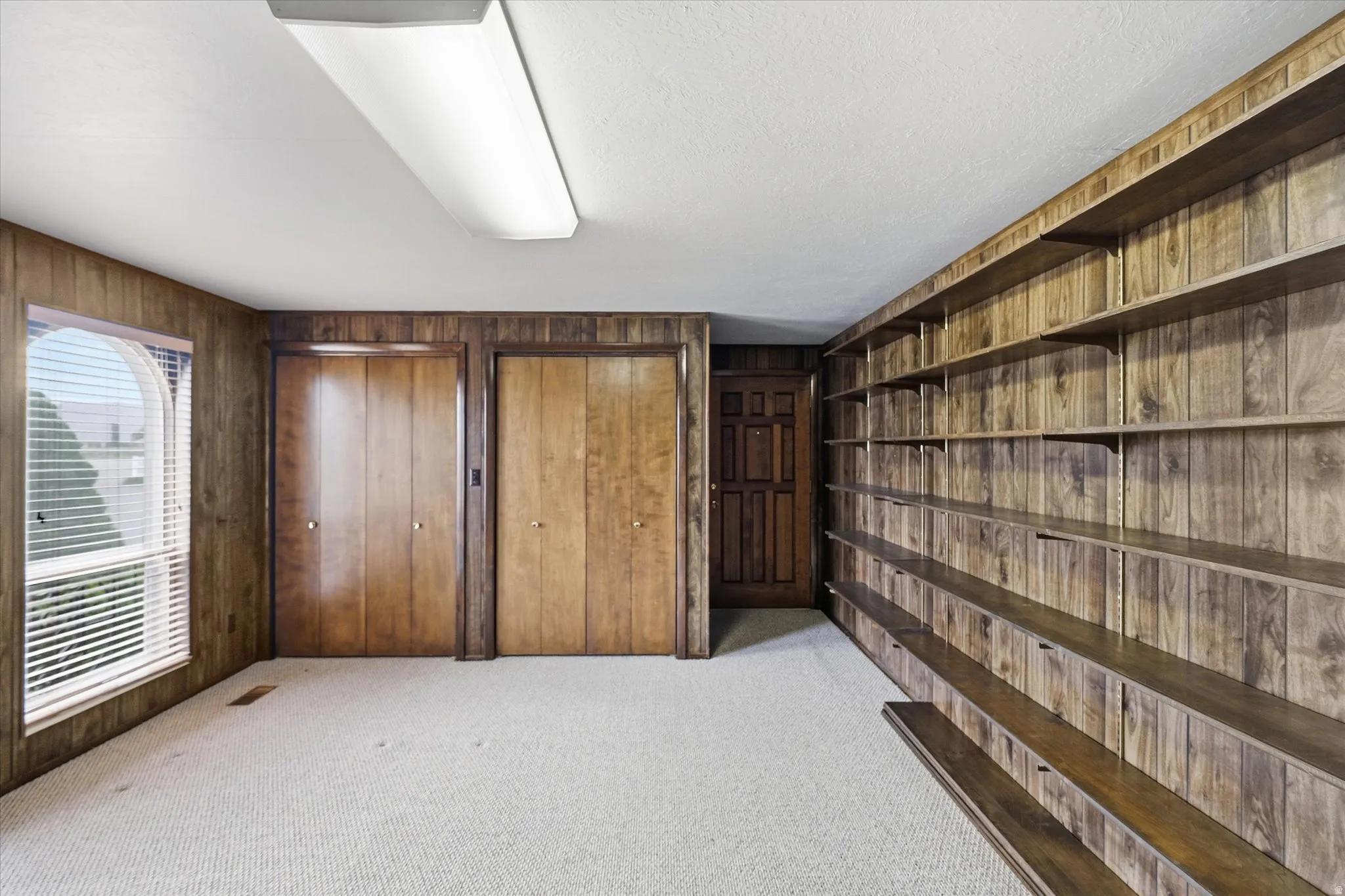 Room featuring wood paneling, extensive built-in shelving, two large closets with wood doors, and a window with blinds