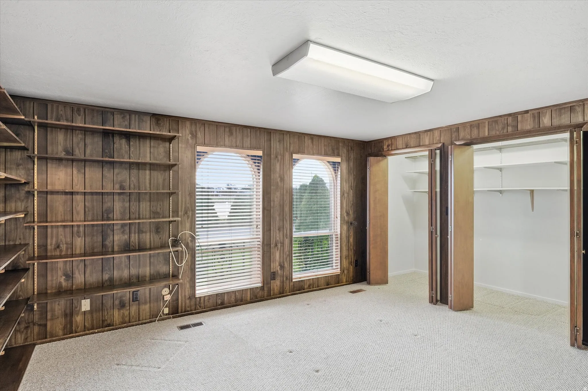 Room featuring wood-paneled walls, built-in shelving, two arched windows with blinds, and white carpeting