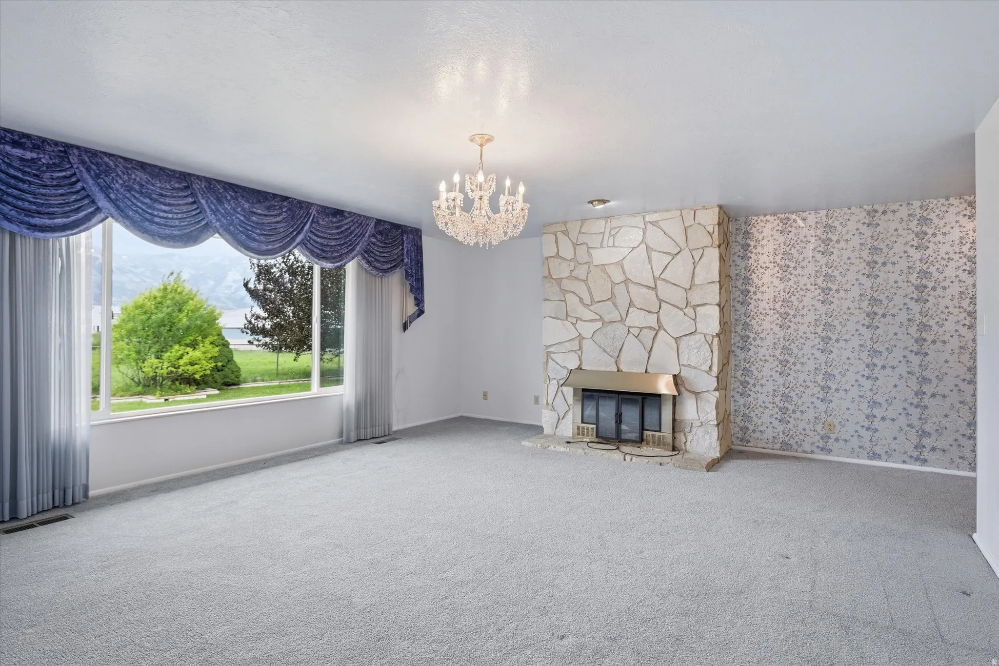 Spacious living area featuring a large picture window, stone-clad fireplace with brass surround, textured accent wall, and a crystal chandelier
