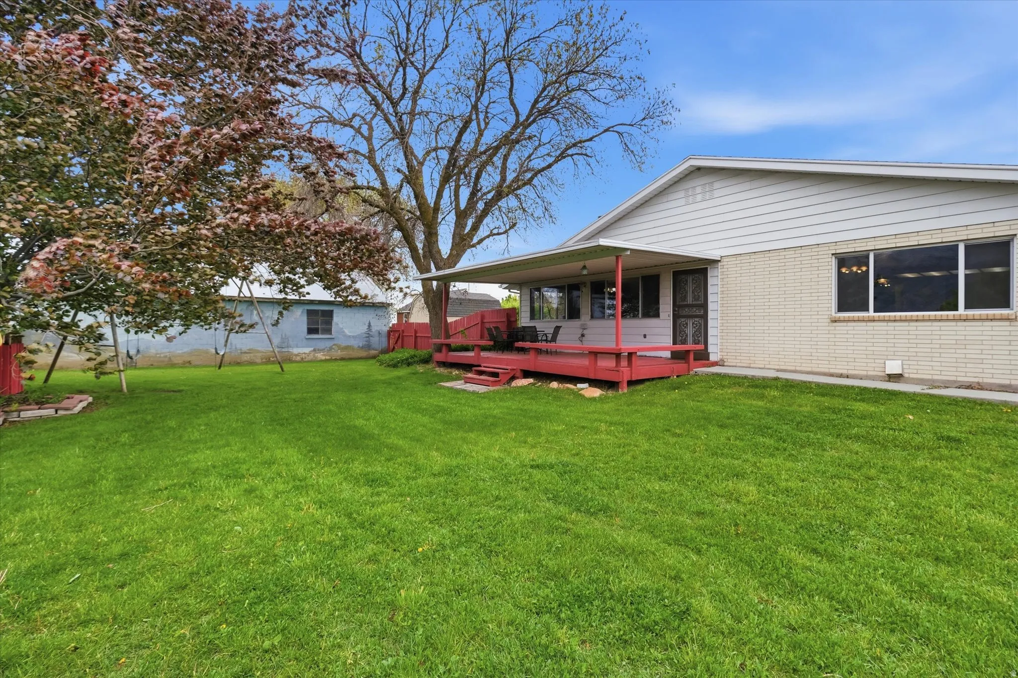 Backyard featuring a vibrant green lawn, mature trees, a red-stained wood deck, and a covered patio area