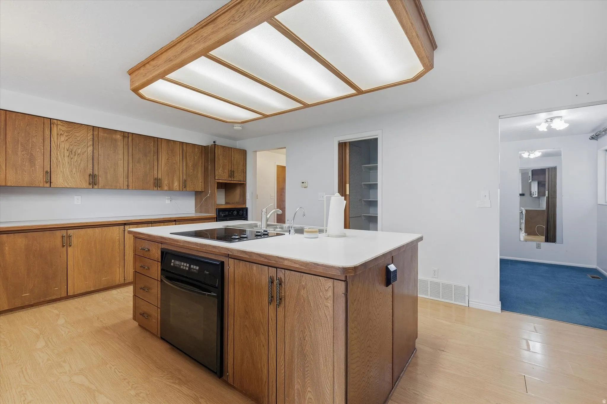 Kitchen featuring wood-finish cabinetry, white countertops, a central island with an integrated cooktop and dishwasher, and light wood-finish flooring