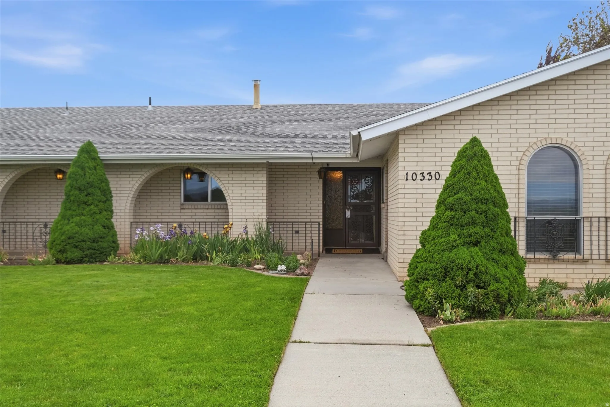 Cream brick exterior with arched porch entries, mature landscaping, a concrete walkway, and an arched window