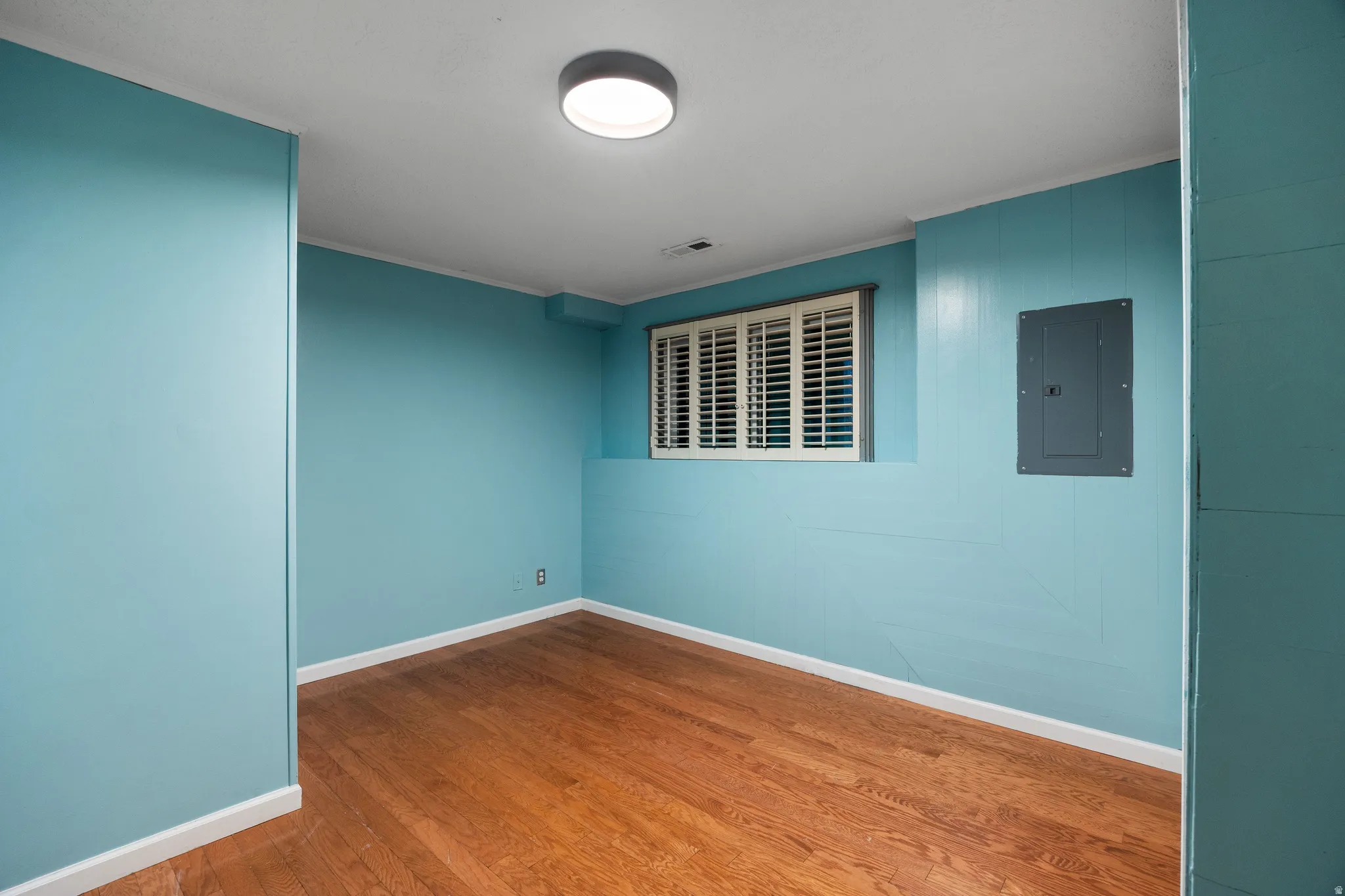 Room featuring wood-finish flooring, light blue paneling, white trim, and a window with white plantation shutters
