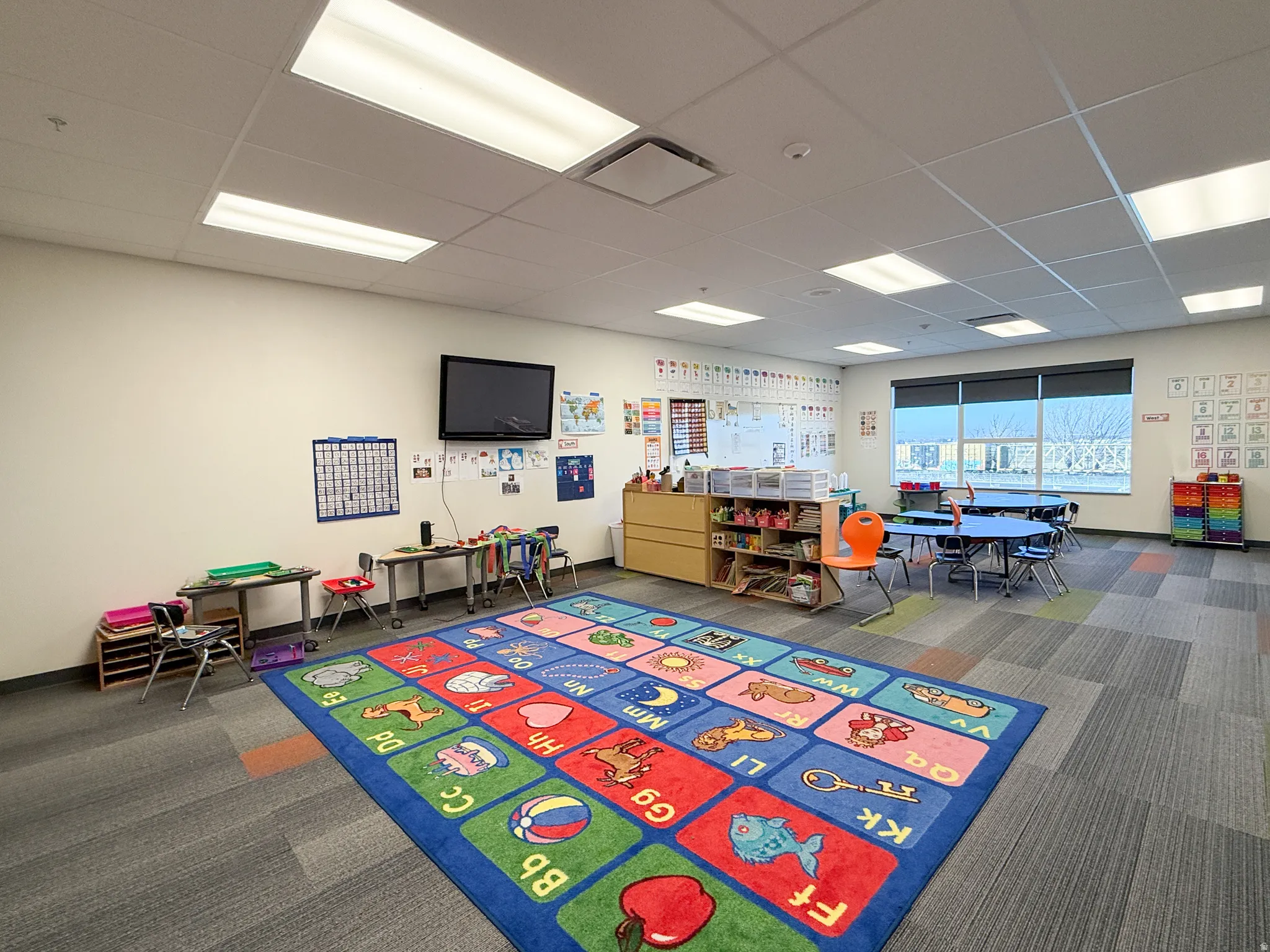 Spacious room featuring modular carpet flooring, ample overhead lighting, and a large window with roller shades
