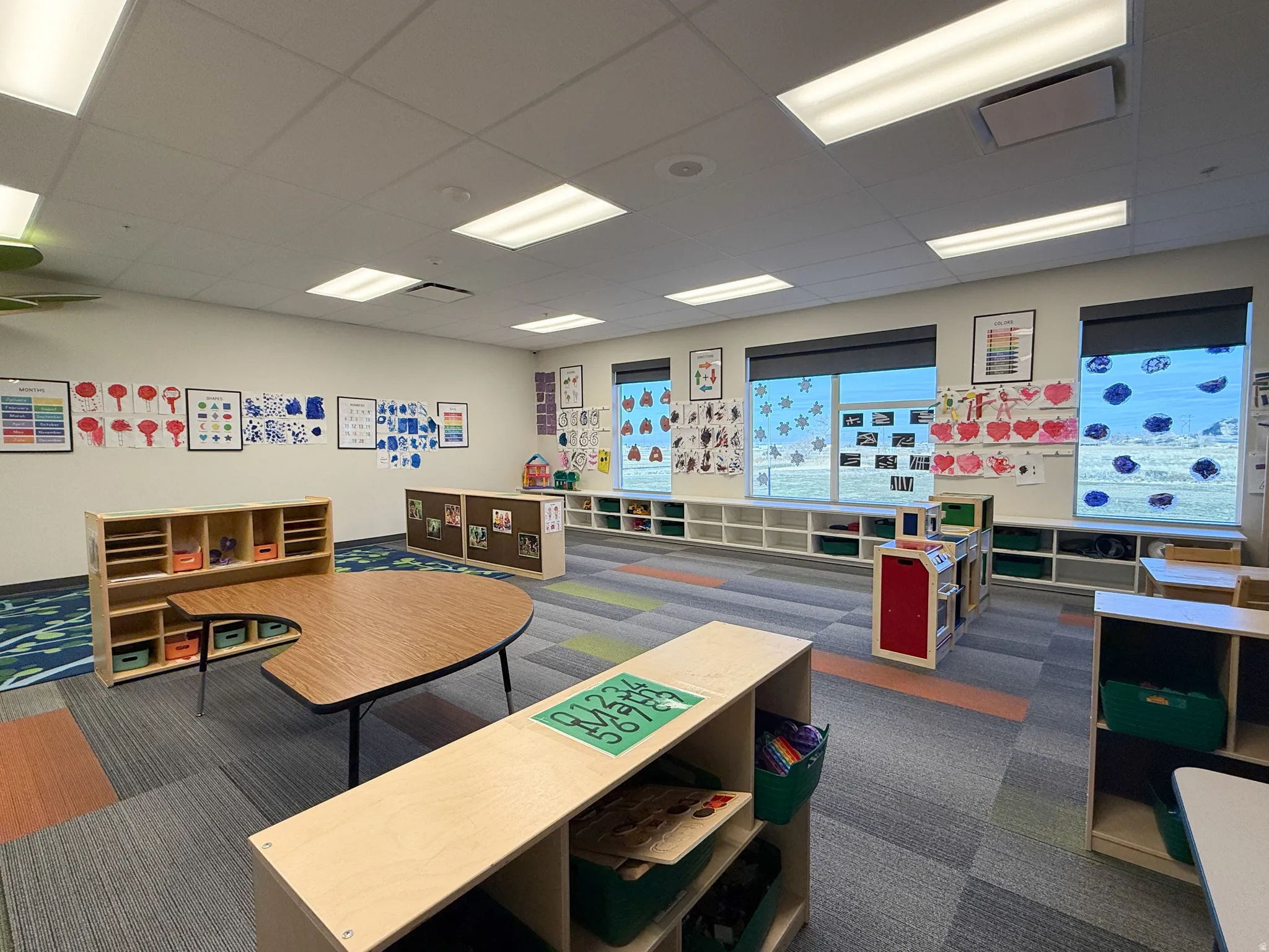 Spacious interior featuring modular carpet tile flooring with color variations, multiple large windows with roller shades, and extensive built-in lower shelving