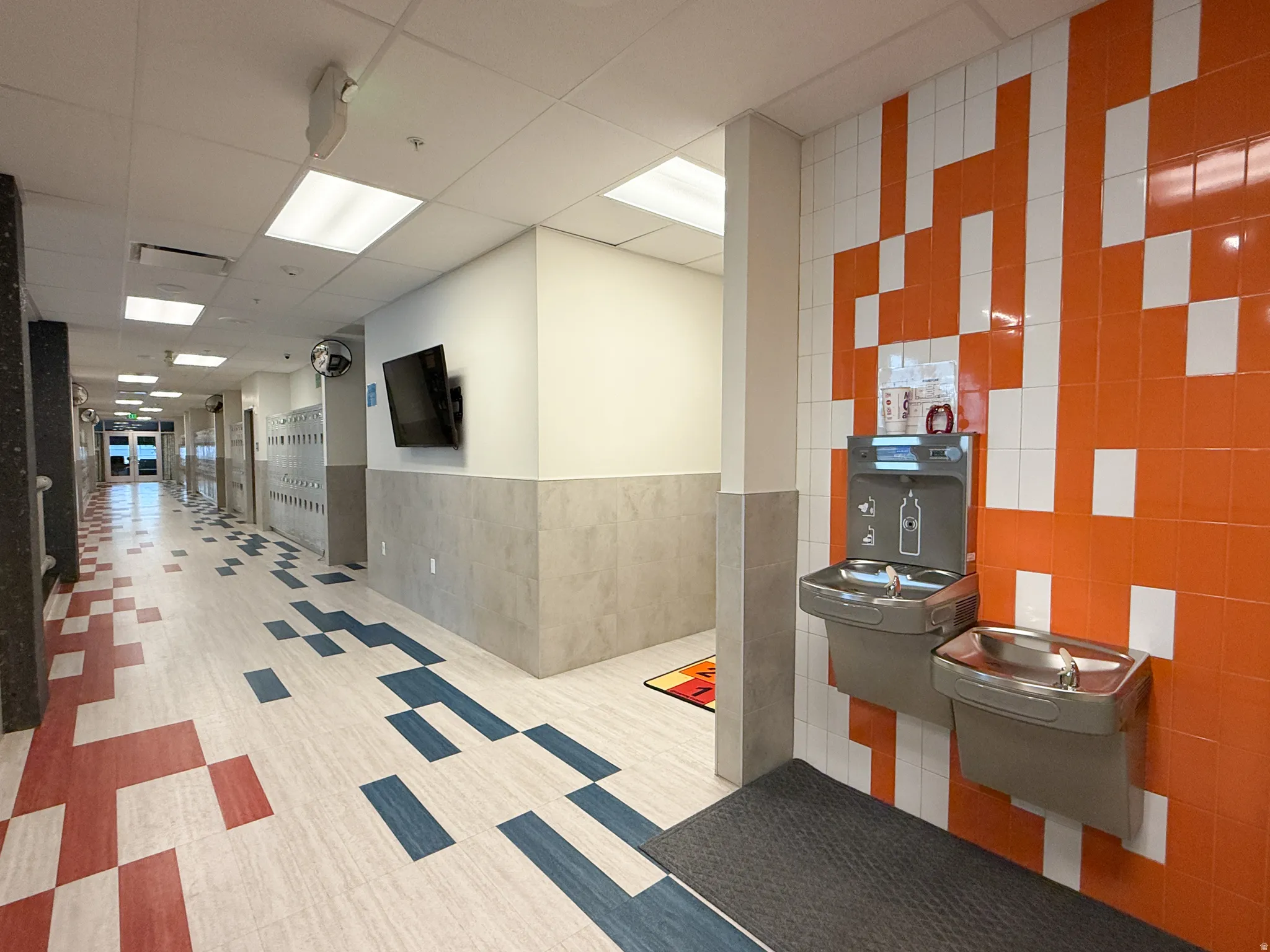 Corridor featuring a water bottle filling station with a traditional drinking fountain, vibrant orange and white patterned wall tile, and light-tone wood-finish flooring with red and blue accents