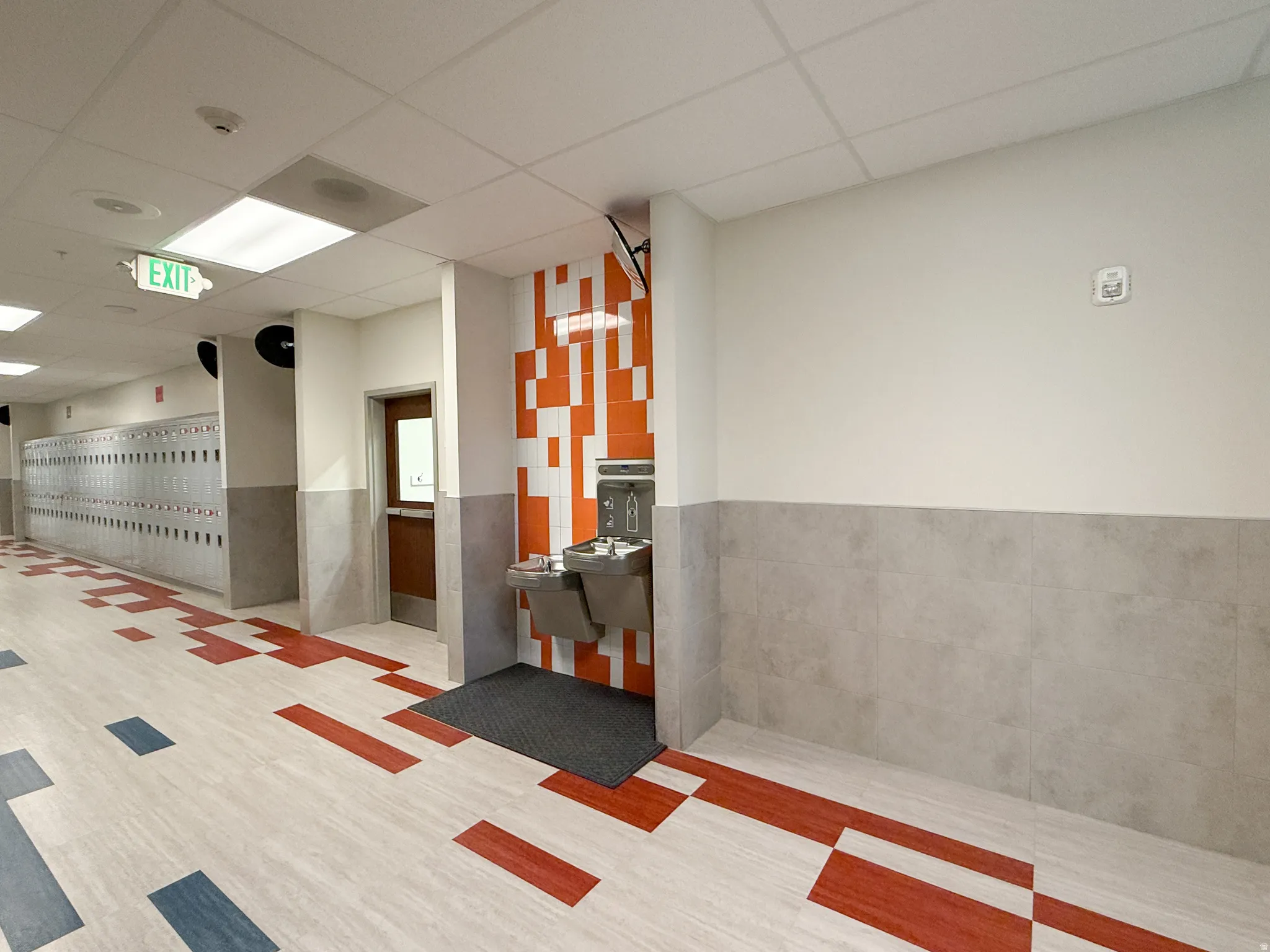 Corridor with light-toned flooring featuring red and blue geometric patterns, complemented by a water fountain station with an orange and white mosaic-tiled accent wall