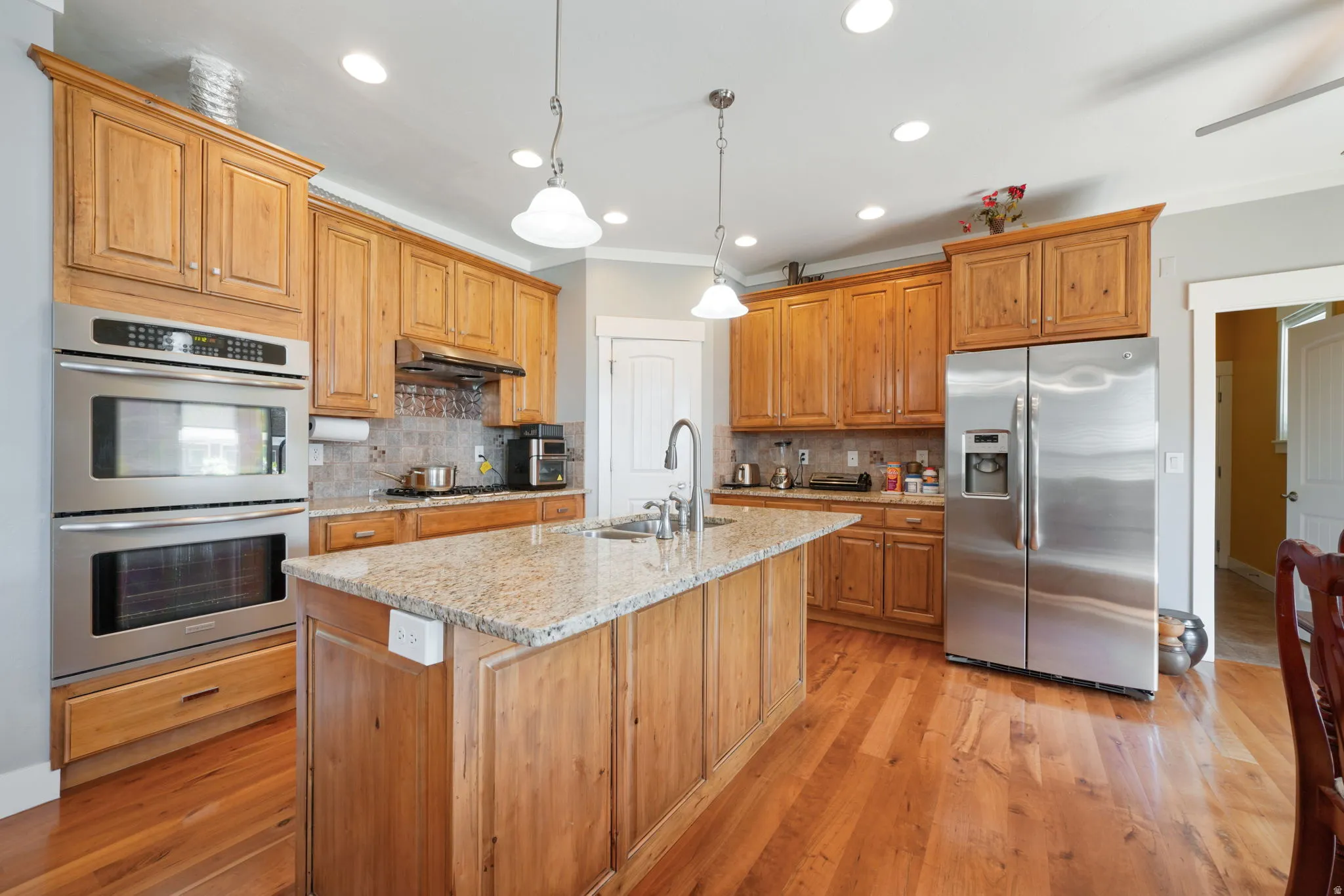 Kitchen featuring wood-finish flooring, a central island with a granite countertop, wood cabinetry, stainless steel double wall ovens, and a stainless steel side-by-side refrigerator