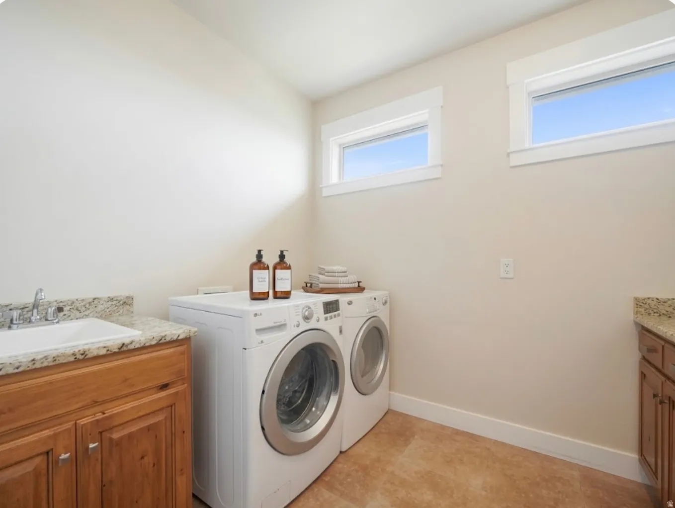 Laundry area featuring light-toned walls, two windows, wood-finish cabinetry, a white basin sink with a chrome faucet, and a light-toned stone-finish countertop