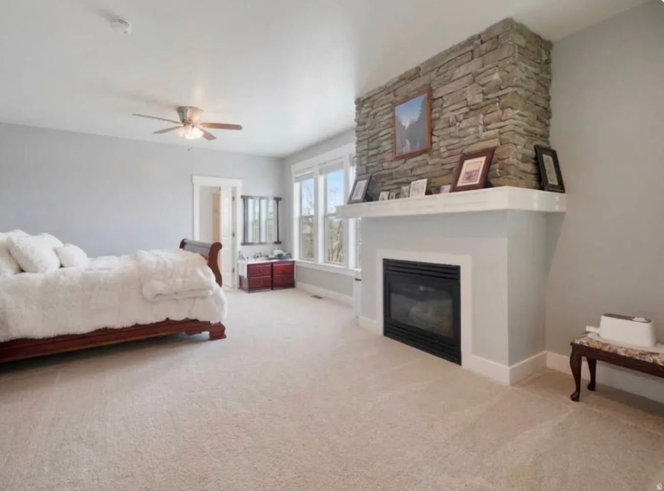 Stone-faced fireplace with a white mantel, ceiling fan with integrated lighting, multiple windows with white trim, and light-toned carpeting