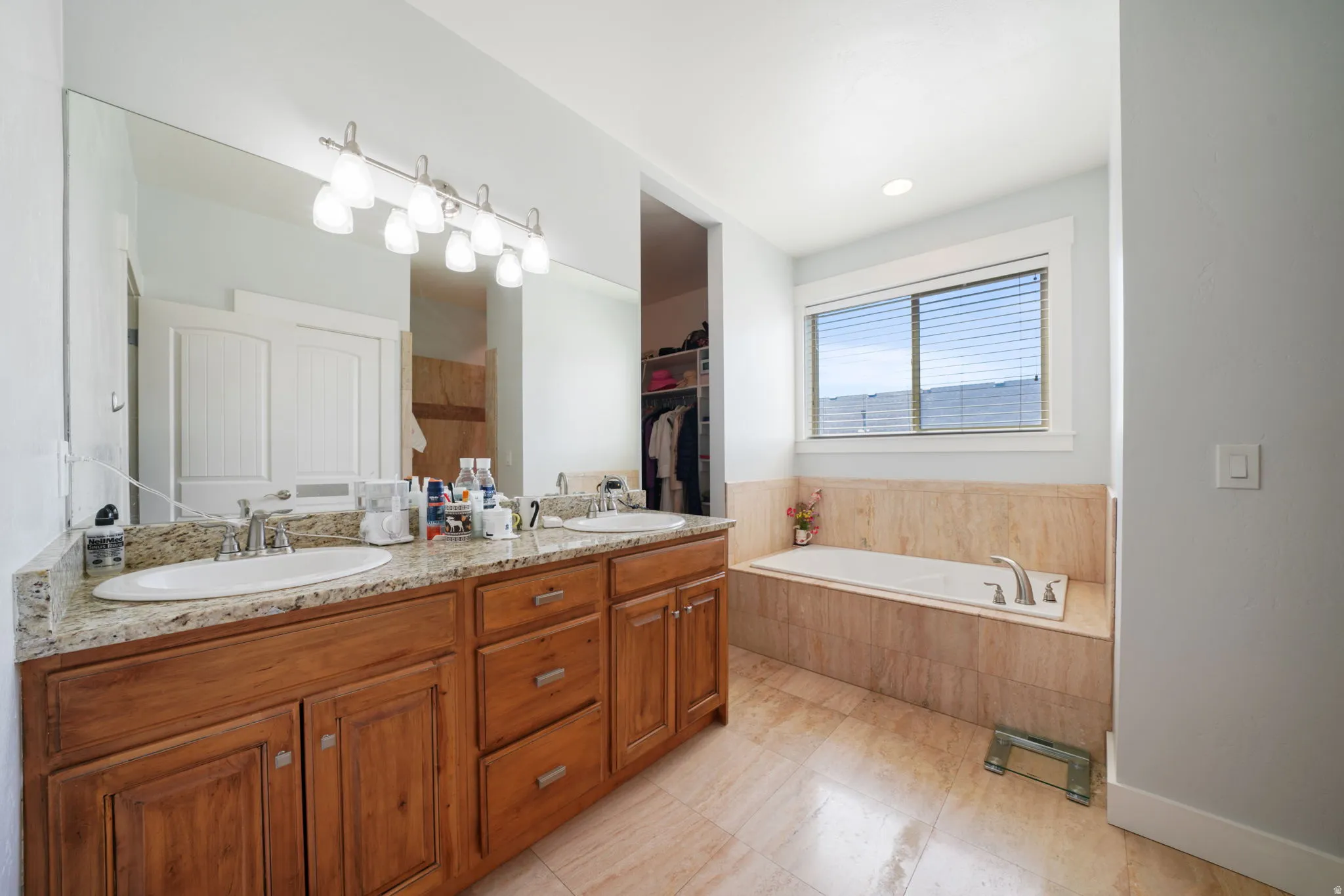 Spacious bathroom featuring a dual vanity with a granite-style countertop, wood cabinetry, a large mirror with overhead lighting, a built-in tub with tile surround, and a window with blinds