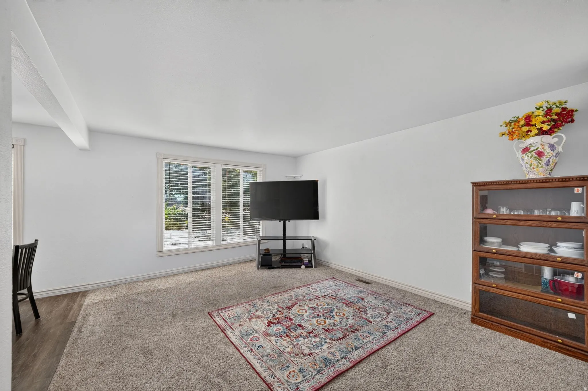 Spacious living area featuring light-toned walls, a large multi-pane window with blinds, and neutral carpeting