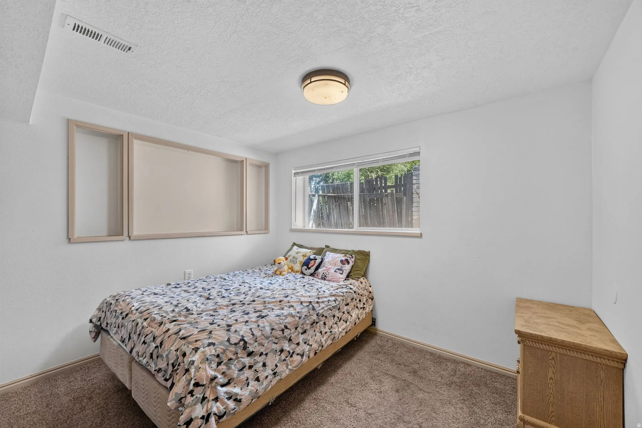 Bedroom featuring a window with exterior fencing visible, neutral carpet flooring, and white painted walls