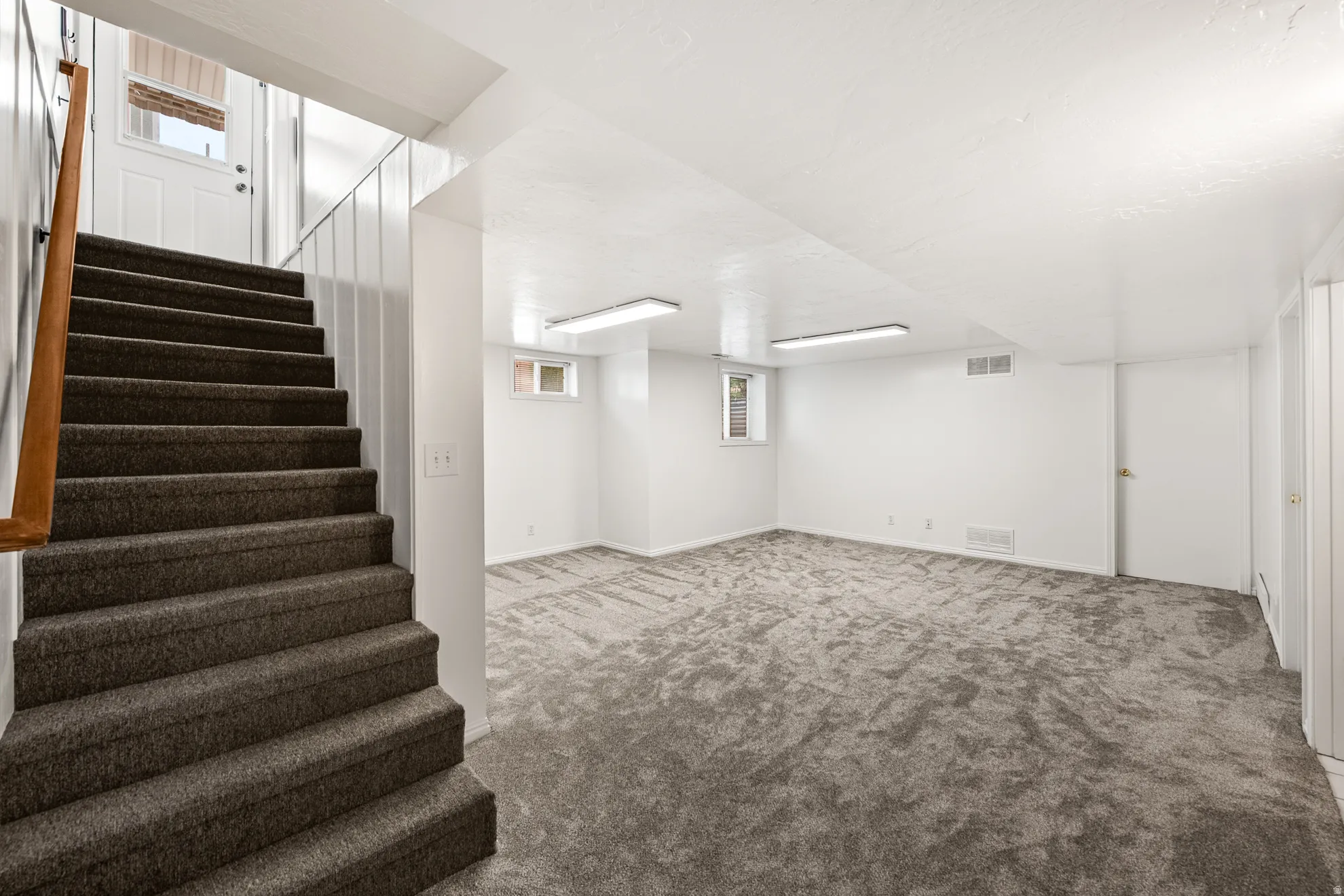 Spacious lower-level room featuring plush gray carpeting, white painted walls, and overhead fluorescent lighting
