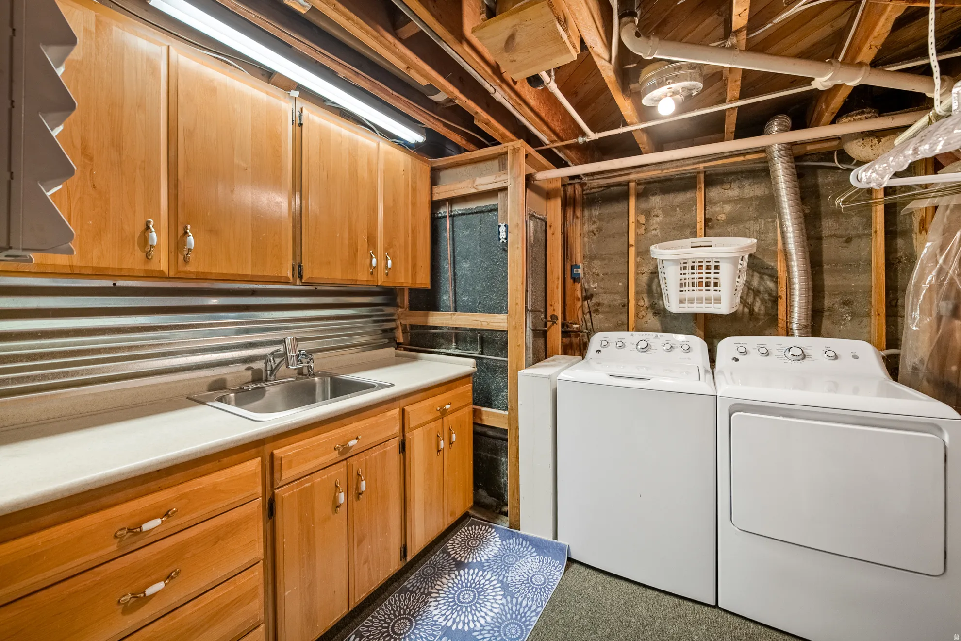 Dedicated laundry space featuring a utility sink, light wood-finish cabinetry, countertop workspace, corrugated metal backsplash, and exposed ceiling joists