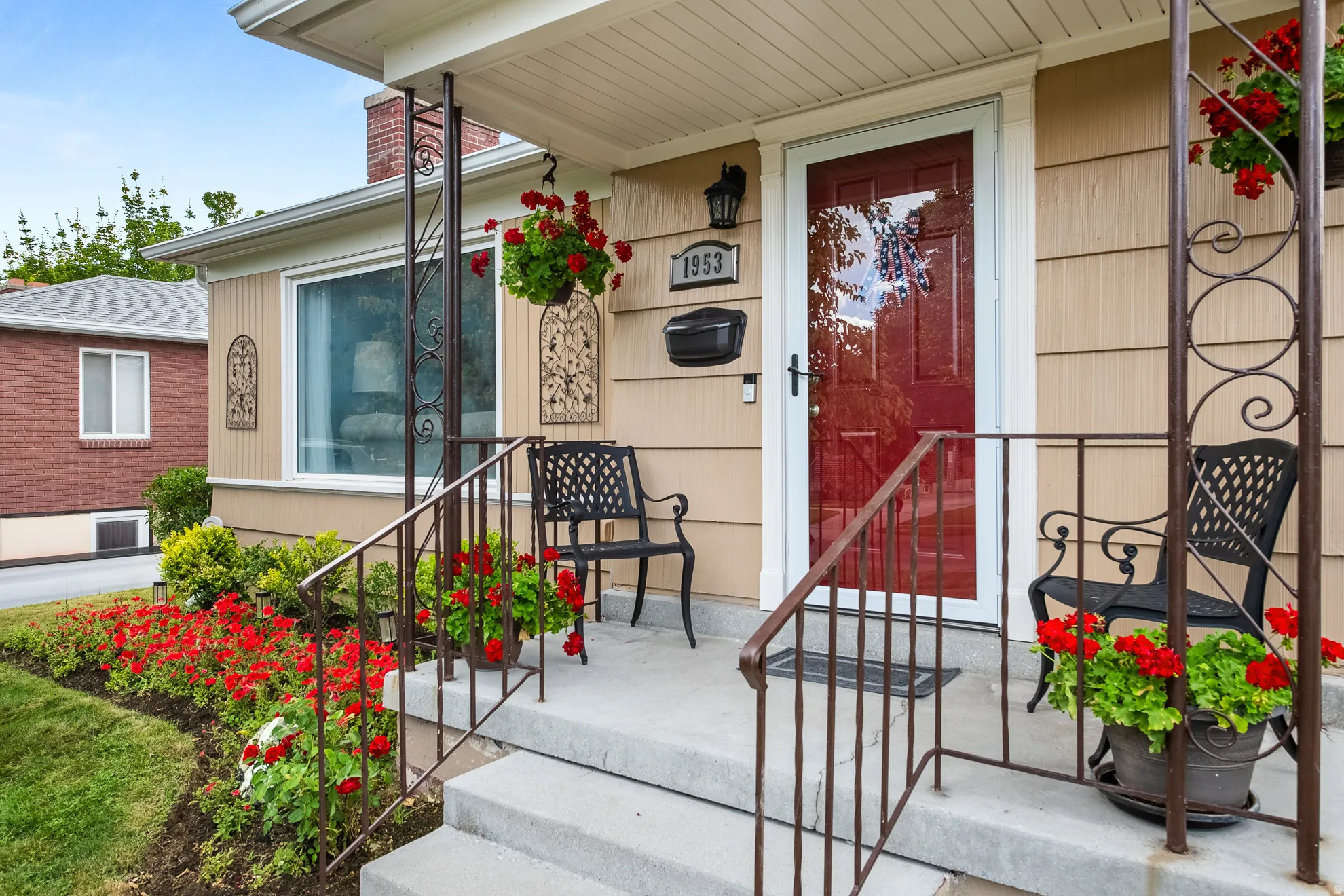 Front porch featuring concrete steps, wrought iron railings, a red entry door, and tan siding