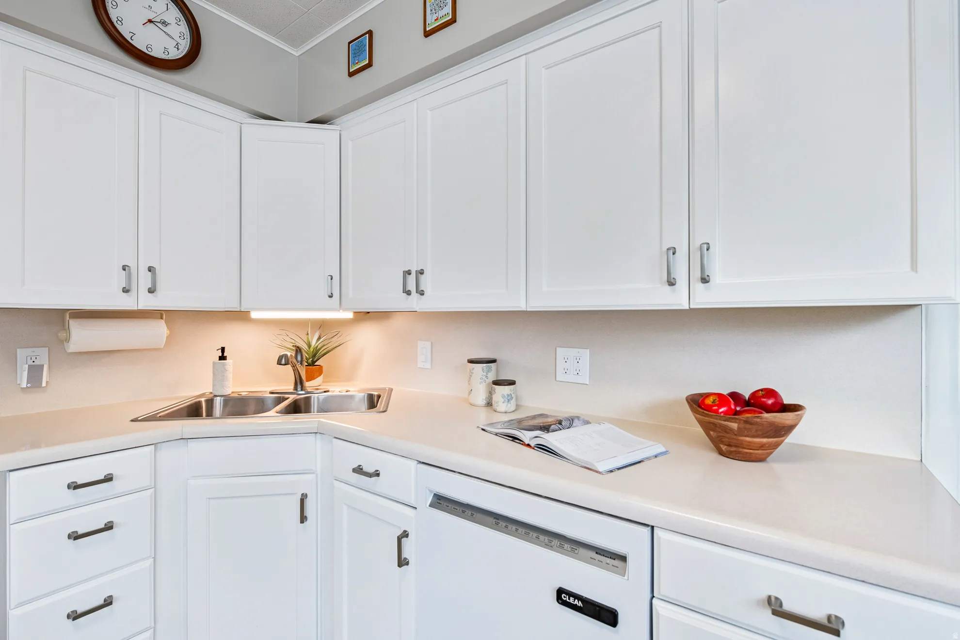 White cabinetry with satin nickel hardware, light-toned countertops, stainless steel double basin sink, and an integrated white dishwasher