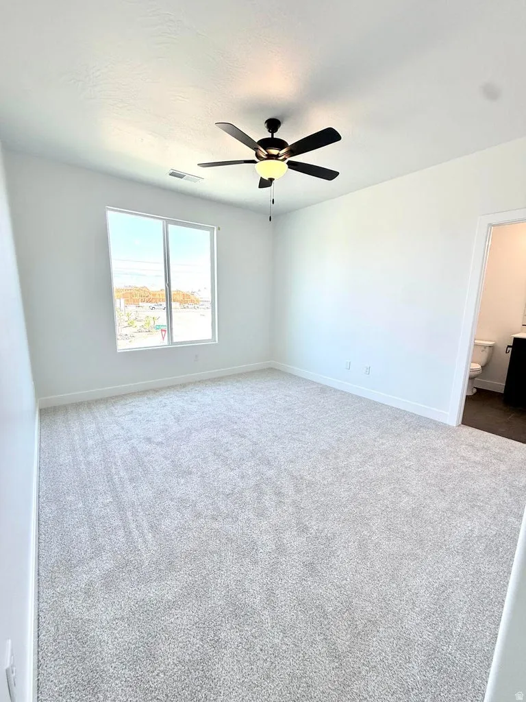 Carpeted room featuring a ceiling fan with integrated lighting, a large window, white baseboards, and a doorway opening to a bathroom with a toilet and vanity