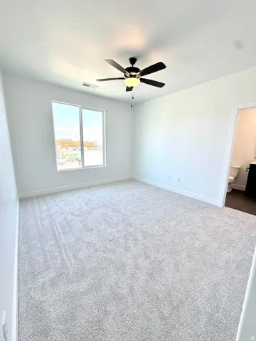 Carpeted room featuring a ceiling fan with integrated lighting, a large window, white baseboards, and a doorway opening to a bathroom with a toilet and vanity