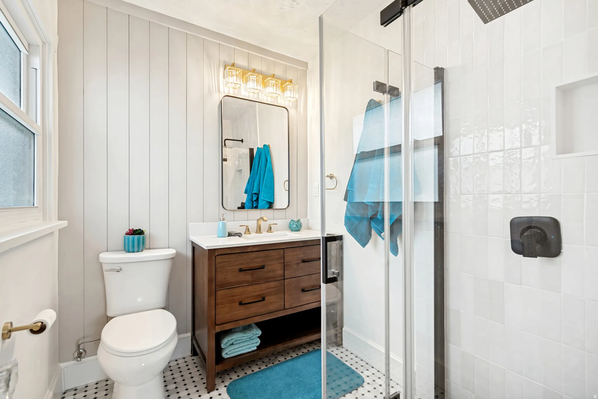 Master/ Primary Bathroom. Bathroom featuring a freestanding wood-finish vanity with a white countertop and gold-tone faucet, a walk-in shower with a glass enclosure and matte black fixtures, shiplap accent wall, and mosaic tile flooring