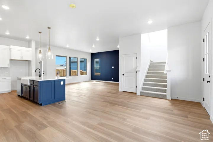 Open-concept living area featuring wood-finish flooring throughout, a modern kitchen with a blue island and white cabinetry, recessed lighting, and a staircase with carpeted treads