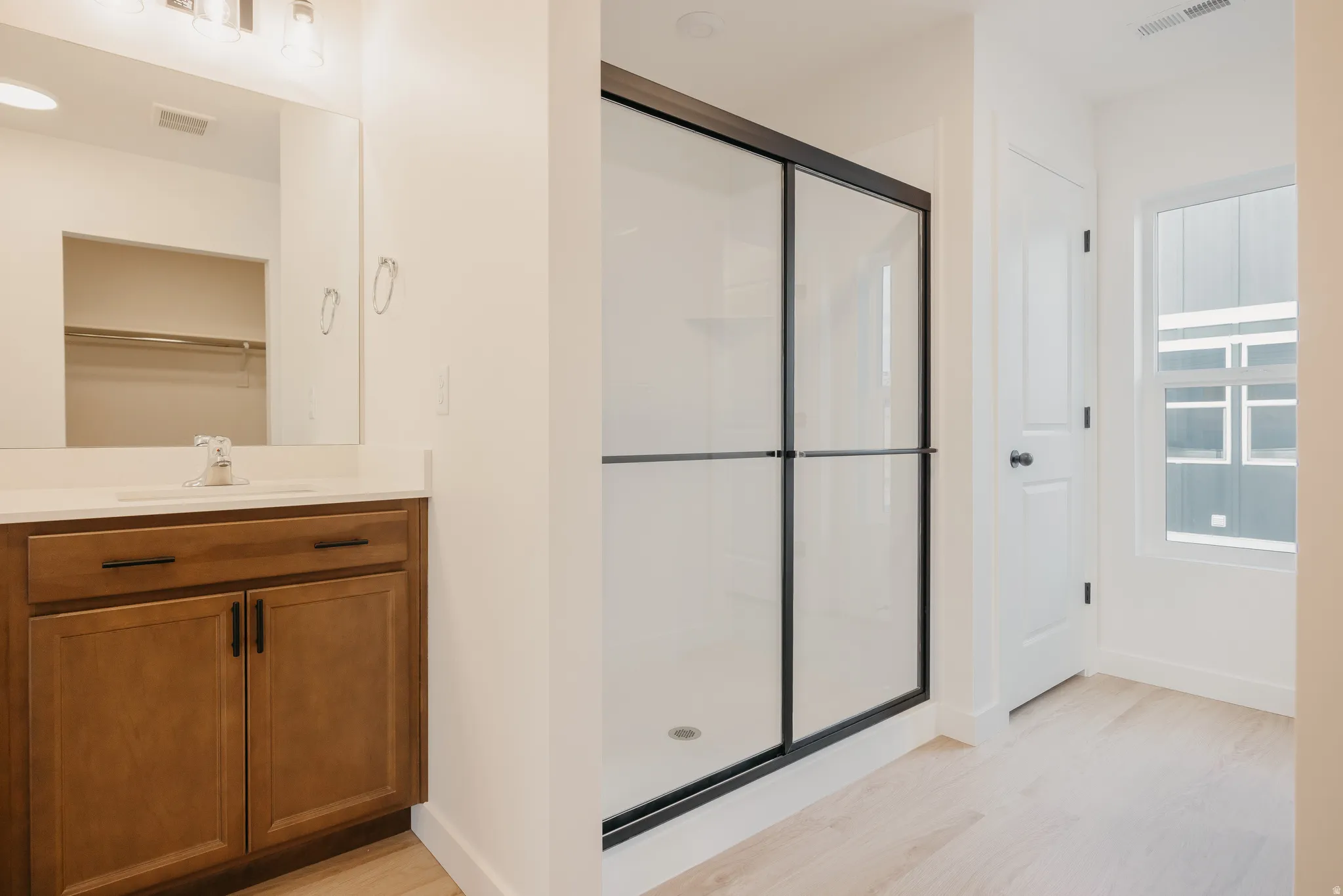 Contemporary bathroom featuring a black-framed glass shower enclosure, a wood-finish vanity with a white countertop and integrated sink, a large wall mirror, and light wood-finish flooring