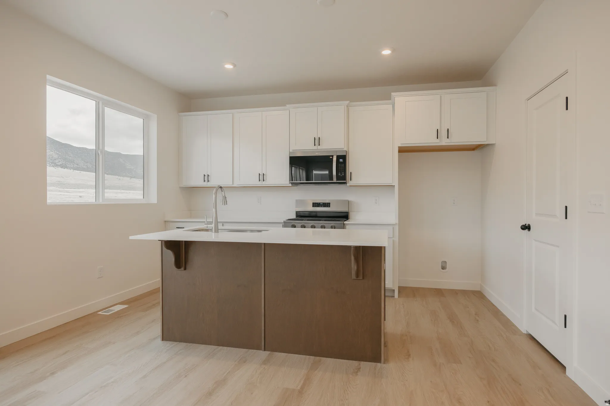 Bright kitchen featuring a central island with an integrated sink, white cabinetry, stainless steel appliances, and light wood-finish flooring